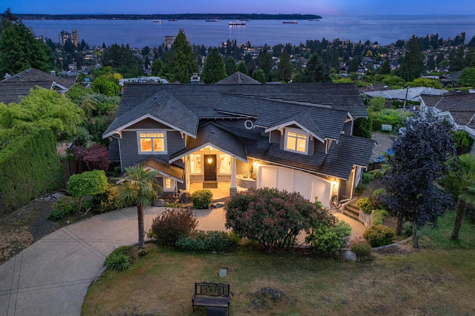 The front of the house from the air at dusk, the drive and the entry lit below the roof