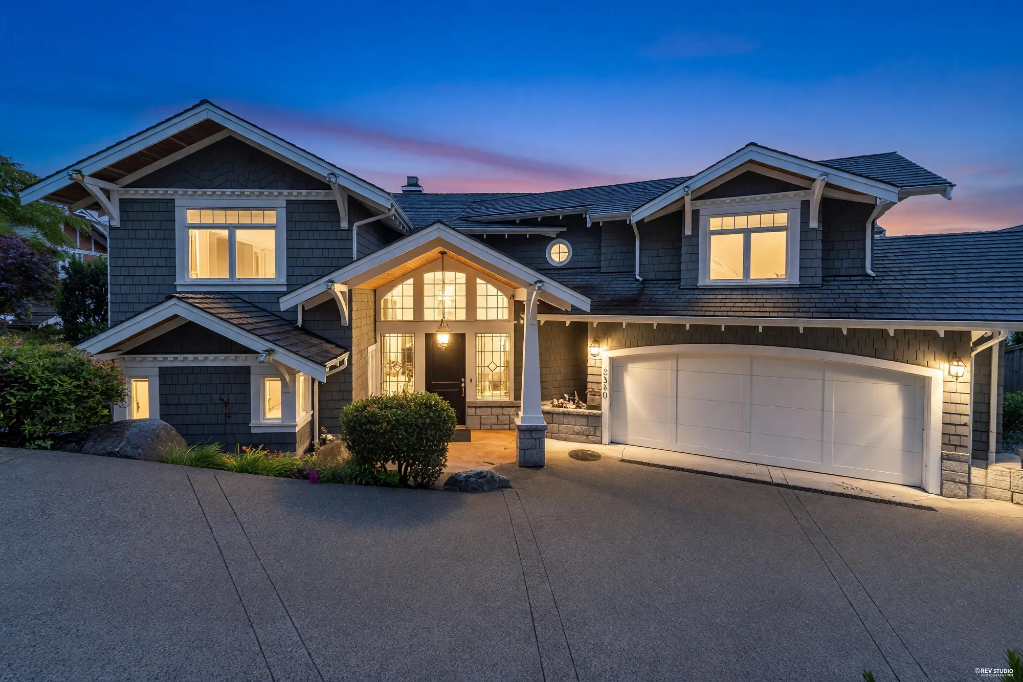 The front of the house at dusk, shingled gables and a lit entry porch above the drive