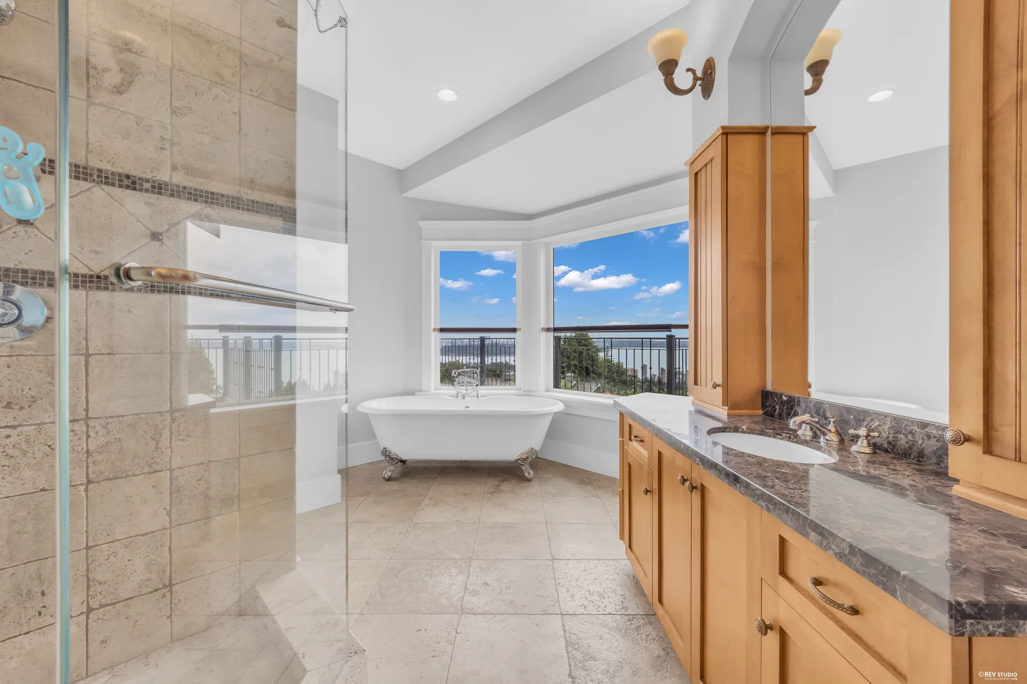The primary bathroom in travertine with a clawfoot tub, a glass shower and twin granite vanities under sconces